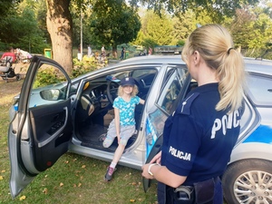 Zdjęcie przedstawia umundurowaną policjantkę, która stoi przy otwartych drzwiach radiowozu, w którym na przednim siedzeniu siedzi dziecko w policyjnej czapce na głowie.