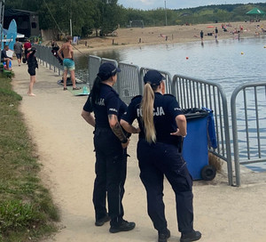 Zdjęcie przedstawia dwie umundurowane policjantki, które stoją przy barierce i wpatrują się w osoby wypoczywające nad zalewem.