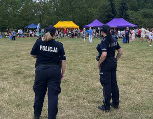 Zdjęcie przedstawia dwie umundurowane policjantki, które stoją na terenie parku i obserwują osoby biorące udział w pikniku.