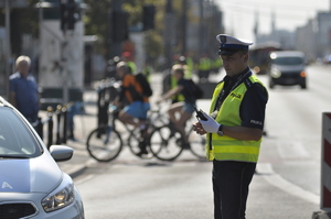 Zdjęcie przedstawia umundurowanego policjanta w odblaskowej kamizelce i białej czapce, który stoi przy pojeździe. Z tyłu, w tle widać sylwetki rowerzystów.