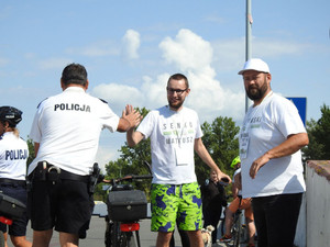 Zdjęcie przedstawia dwóch mężczyzn ubranych w jasne koszulki, którzy stoją przodem do obiektywu aparatu. Jeden z nich przybija &quot;piątkę&quot; z jadącym na rowerze policjantem ubranym w strój sportowy.