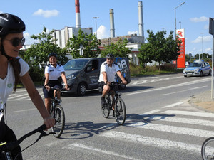 Zdjęcie przedstawia jadących na rowerach policjantów ubranych w strój sportowy, tj. jasne koszulki, ciemne spodenki i ciemne kaski rowerowe.