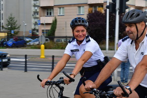 Zdjęcie przedstawia jadących na rowerach policjantów ubranych w strój sportowy, tj. jasne koszulki, ciemne spodenki i ciemne kaski rowerowe.