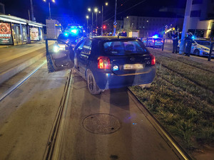 Zdjęcie przedstawia samochód osobowy w kolorze ciemnym, który stoi prawymi kołami na torowisku tramwajowym. Przed nim widać oznakowany radiowóz z włączonymi światłami na dachu.