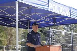 Zdjęcie przedstawia umundurowanego policjanta stojącego przy mikrofonie pod niebieskim zadaszeniem.