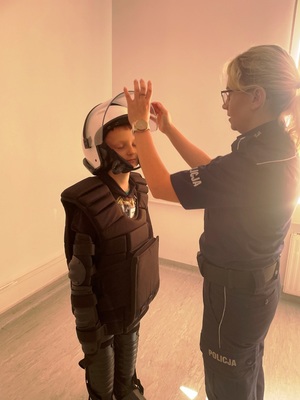 Zdjęcie przedstawia policjantkę stojącą przed małym chłopcem, który ubrany jest w elementy policyjnego stroju wykorzystywanego przy zabezpieczeniach. Policjantka pomaga mu odpowiednio nałożyć policyjny kask na głowę.
