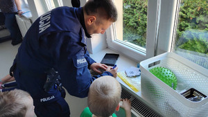Zdjęcie przedstawia umundurowanego policjanta, który pochyla się nad parapetem.Przy nim widać małego chłopca.