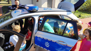 Zdjęcie przedstawia umundurowanego policjanta, który stoi przy radiowozie, wewnątrz którego siedzi dużo dzieci.