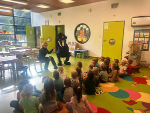 Zdjęcie przedstawia umundurowaną policjantkę, która siedzi obok postaci przebranej w postać maskotki Borsuka. Policjantka ma uniesioną do góry prawą rękę. Przed nimi na dywanie siedzą dzieci.