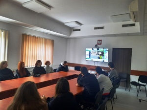 Zdjęcie przedstawia uczniów siedzących wokół stołu i patrzących w ekran telewizora umieszczonego na ścianie.