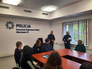 Zdjęcie przedstawia uczniów siedzących wokół stołu i patrzących w stronę stojących policjantów.