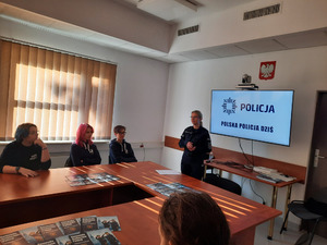 Zdjęcie przedstawia umundurowaną policjantkę, która stoi przed młodzieżą siedzącą przy stole. Przy policjantce widać ekran telewizora, na którym wyświetla się napis POLSKA POLICJA.