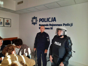 Zdjęcie przedstawia umundurowanego policjanta, który stoi przy osobie ubranej w umundurowanie policjanta na zabezpieczenie imprezy masowej. Obok nich widać osoby siedzące przy stole.