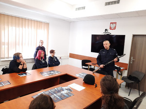 Zdjęcie przedstawia umundurowanego policjanta, który stoi przed osobami siedzącymi przy stole i trzyma w dłoniach strzelbę.