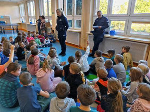 Zdjęcie przedstawia umundurowanych policjantów, którzy stoją przed dziećmi siedzącymi na dywanie.