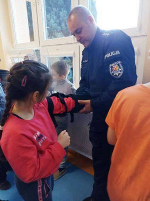 Zdjęcie przedstawia umundurowanego policjanta, który zakłada na rękę czarny element umundurowania dziewczynce stojącej obok.