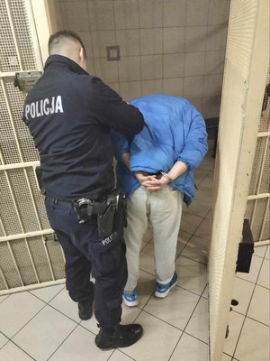 Zdjęcie przedstawia umundurowanego policjanta, który prowadzi po swojej prawej stornie zatrzymanego mężczyznę. Mężczyzna ten jest pochylony do przodu, Na rękach trzymanych z tyłu ma założone kajdanki, Jest ubrany w niebieską kurtkę puchową i jasnoszare spodnie dresowe. Przed nim widać otwarte okratowane drzwi.