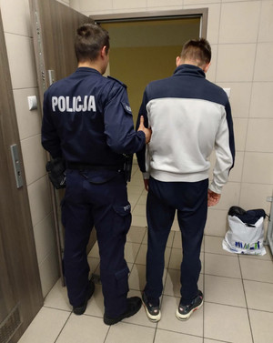 Zdjęcie przedstawia umundurowanego policjanta, który prowadzi po swojej prawej stronie zatrzymanego mężczyznę. Obaj stoją tyłem do zdjęcia. Zatrzymany mężczyzna ubrany jest w bluzę w kolorze biało-granatowym i ciemne spodnie. Mężczyzna ma pochyloną do dołu głowę.  Przed nimi przy ścianie stoi na podłodze jasna reklamówka.