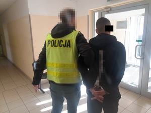 Zdjęcie przedstawia policjanta ubranego w kurtkę i odblaskową kamizelkę z napisem POLICJA, który prowadzi po swojej prawej stronie zatrzymanego mężczyznę. Twarz policjanta jest zasłonięta komputerowym filtrem. Zatrzymany mężczyzna ma kajdanki założone na rękach trzymanych z tyłu. Jest ubrany na ciemno. Część twarzy zasłania mu czarny prostokąt. Mężczyźni stoją na korytarzu, kierując się do wyjścia z budynku.