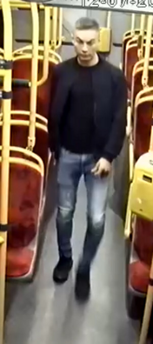 Zdjęcie przedstawia mężczyznę stojącego wewnątrz autobusu. Jest on skierowany frontem do kamery monitoringu. Ubrany jest w ciemną kurtkę i bluzkę, ciemnogranatowe spodnie i ciemne buty.