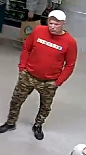 Zdjęcie przedstawia mężczyznę ubranego w białą czapkę z daszkiem, czerwoną bluzkę z białym napisem na wysokości klatki piersiowej i wzorzyste spodnie w odcieniu zielonym. Mężczyzna stoi przodem do kamery monitoringu. Trzyma dłonie w kieszeniach spodni.