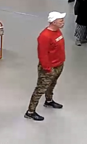 Zdjęcie przedstawia mężczyznę ubranego w białą czapkę z daszkiem, czerwoną bluzkę z białym napisem na wysokości klatki piersiowej i wzorzyste spodnie w odcieniu zielonym. Mężczyzna stoi prawym profilem do kamery monitoringu. Trzyma dłonie w kieszeniach spodni.