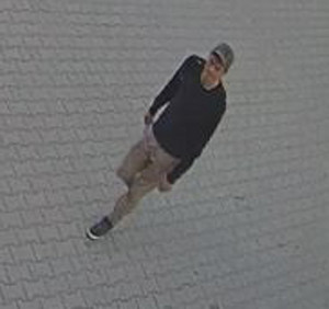 Zdjęcie przedstawia mężczyznę idącego chodnikiem. Jest on ubrany w ciemną bluzę, beżowe krótkie spodenki, ciemną czapkę i ciemne buty.