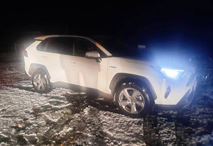 Zdjęcie przedstawia biały zaparkowany na działce z włączonymi światłami samochód.