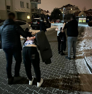Zdjęcie przedstawia nieumundurowanych policjantów, którzy prowadzą po ulicy dwóch pochylonych mężczyzn, którzy mają kajdanki założone na ręce trzymane z tyłu. Zatrzymani mężczyźni są ubrani w jasne koszulki na krótki rękaw. W tle widać budynek mieszkalny i zaparkowane pojazdy.