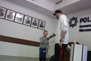 Zdjęcie przedstawia mężczyznę stojącego w białej bluzie i czapce z daszkiem na głowie, który podaje rękę chłopcu stojącemu po jego prawej stronie.