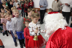 Zdjęcie przedstawia dziewczynkę stojącą przed Mikołajem, która trzyma w dłoniach złoty Mikołajowy dzwonek.