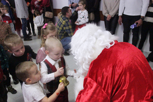 Zdjęcie przedstawia małego chłopca stojącego przed Mikołajem, który trzyma w dłoniach złoty Mikołajowy dzwonek.