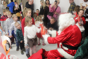 Zdjęcie przedstawia dziewczynkę stojącą przed Mikołajem, która odbiera od Mikołaja torebkę z prezentem.
