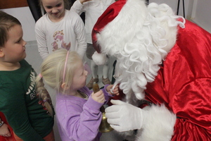 Zdjęcie przedstawia dziewczynkę stojącą przed Mikołajem, która trzyma w dłoni złoty dzwonek.