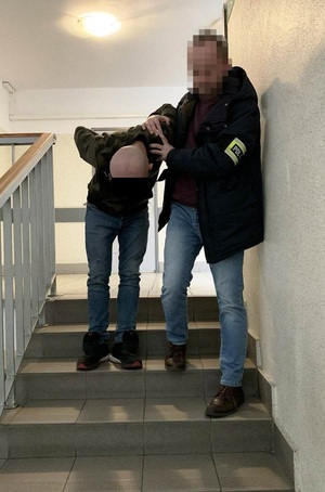Zdjęcie przedstawia policjanta ubranego w ciemną kurtkę i niebieskie spodnie jeansowe oraz odblaskową opaskę na lewym przedramieniu z napisem POLICJA, który sprowadza po schodach wewnątrz budynku zatrzymanego mężczyznę. Mężczyzna idzie zgięty w pół. Ma pochyloną głowę. Część twarzy zasłania mu czarny prostokąt. Mężczyzna jest ubrany w ciemną kurtkę i niebieskie spodnie jeansowe.