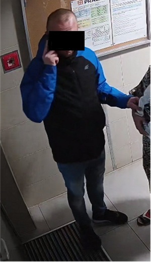 Zdjęcie przedstawia mężczyznę ubranego w niebieską kurtkę, który stoi na klatce schodowej. Mężczyzna trzyma na wysokości prawego policzka dłoń. Część jego twarzy zasłania czarny prostokąt. Po prawej stronie zdjęcia widać fragment sylwetki innej osoby.