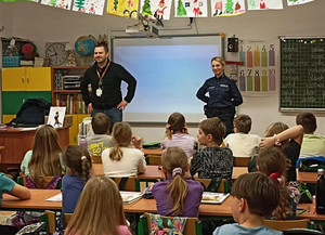 Zdjęcie przedstawia umundurowaną policjantkę i mężczyznę ubranego na ciemno, którzy stoją przed dziećmi siedzącymi w ławkach szkolnych.