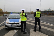 Zdjęcie przedstawia dwoje policjantów ubranych w mundury i odblaskowe kamizelki, którzy stoją przy oznakowanym radiowozie zaparkowanym wzdłuż drogi.
