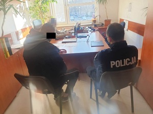 Zdjęcie przedstawia umundurowanego policjantka, który siedzi po prawej stronie od zatrzymanego mężczyzny. Część twarzy mężczyzny zasłania czarny prostokąt. Przy biurku pod oknem siedzi policjant, jego twarz jest zasłonięta komputerowym filtrem. Na biurku leżą rozłożone dokumenty.