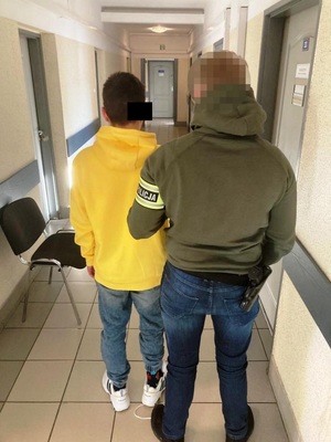 Zdjęcie przedstawia policjanta ubranego w ciemną kurtkę z seledynową opaską na lewym przedramieniu z napisem POLICJA, granatowe spodnie jeansowe. Jego twarz jest zasłonięta komputerowym filtrem. Po jego lewej stronie stoi zatrzymany mężczyzna. Ma on na sobie długą żółtą bluzę, granatowe spodnie jeansowe i białe buty sportowe. Część jego twarzy zasłania czarny prostokąt. Obaj stoją tyłem do obiektywu aparatu.