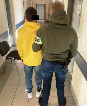 Zdjęcie przedstawia policjanta ubranego w ciemną kurtkę z seledynową opaską na lewym przedramieniu z napisem POLICJA, granatowe spodnie jeansowe. Jego twarz jest zasłonięta komputerowym filtrem. Po jego lewej stronie stoi zatrzymany mężczyzna. Ma on na sobie długą żółtą bluzę, granatowe spodnie jeansowe i białe buty sportowe. Część jego twarzy zasłania czarny prostokąt. Obaj stoją tyłem do obiektywu aparatu.