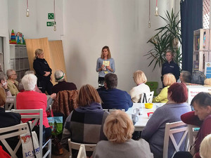 Zdjęcie przedstawia dwie policjantki i kobietę stojące przed siedzącymi na krzesłach seniorami.