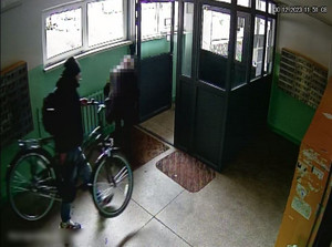Zdjęcie przedstawia ubranego na ciemno mężczyznę niosącego rower. W tle widać postać osoby stojącej tyłem.