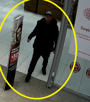 Zdjęcie przedstawia mężczyznę wchodzącego do sklepu. Ma około 30 lat, około 170 cm wzrostu, normalną budowę ciała, ciemny zarost na twarzy. Ubrany jest w czarną czapkę z daszkiem z białym napisem „COOL” z przodu, czarną błyszczącą kurtkę pikowaną, czarny sweter, czarne spodnie i czarne zimowe buty za kostkę. Po obu jego stronach widać kolorowe banery reklamowe.
