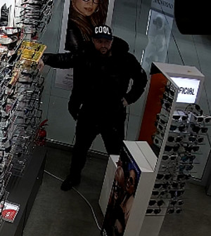Zdjęcie przedstawia mężczyznę stojącego w sklepie pomiędzy półkami z okularami. Ma on około 30 lat, około 170 cm wzrostu, normalną budowę ciała, ciemny zarost na twarzy. Ubrany jest w czarną czapkę z daszkiem z białym napisem „COOL” z przodu, czarną błyszczącą kurtkę pikowaną, czarny sweter, czarne spodnie i czarne zimowe buty za kostkę.