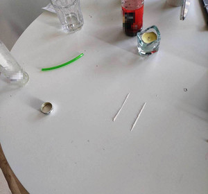 Zdjęcie przedstawia butelki i inne przedmioty ustawione na białym blacie stołu. Na środku widać dwie równolegle usypane kreski białego proszku.
