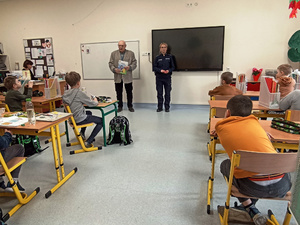 Zdjęcie przedstawia umundurowaną policjantkę, która stoi obok mężczyzny trzymającego w rękach kolorową książeczkę. Oboje stoją przed uczniami siedzącymi w ławkach.