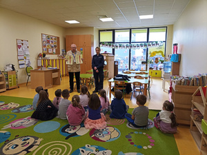 Zdjęcie przedstawia umundurowaną policjantkę oraz mężczyznę trzymającego w dłoniach kolorowe książeczki edukacyjne, którzy stoją przed dziećmi siedzącymi na dywanie.