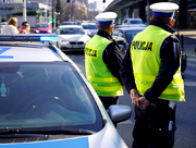 Zdjęcie przedstawia dwóch policjantów ubranych w odblaskowe kamizelki, którzy stoją przy zaparkowanym radiowozie i wpatrują się w jadące samochody.