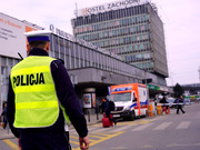 Zdjęcie przedstawia umundurowanego policjanta ubranego w odblaskową kamizelkę i granatowo-białą czapkę, który stoi przy ulicy. Przed nim widać autobus i wysoki budynek.